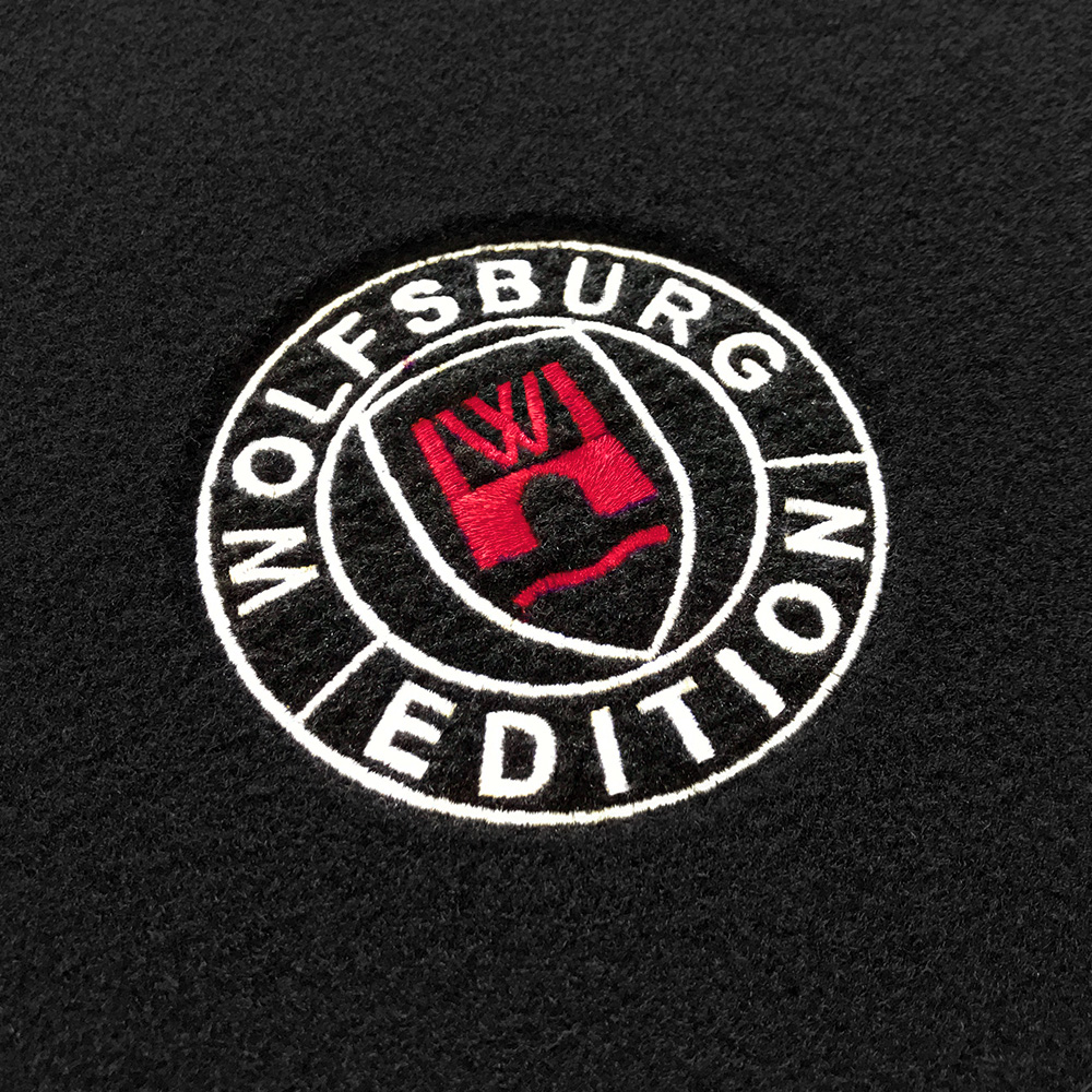 Das gestickte Wolfsburg Editions Logo auf einem schwarzen Teppich und das Logo ist in rot.